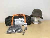 Lightweight dome tent in green carry bag with orange straps, Windriver bucket hat on mannequin head, wireless headphones in plastic and metal camping spoon on table.