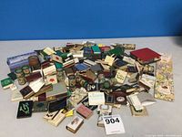 Wide view of a large assortment of vintage match boxes and holders arranged on a flat surface showing diversity in size, shape, and design.