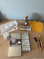 Full view of painting set including box, paint tubes, brushes, stencils, palette, ruler, and paint bowl on tabletop.