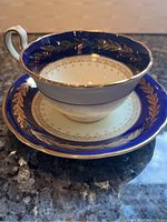 Side view of the Aynsley bone china teacup on its matching saucer displaying the cobalt blue and gold leaf decorative borders.