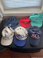 Seven assorted ball caps in various colors and brands arranged on a black surface under a window blind.