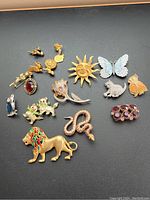 Photo showing a collection of 17 lapel pin brooches arranged on a flat surface, depicting diverse designs including animals, flowers, and symbolic motifs in gold-tone metal with enamel and rhinestones.