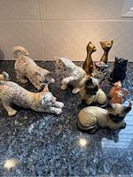 Photo of 11 ceramic and brass cat figurines arranged on a granite countertop. Includes floral pattern cats, brass statues, and painted realistic cats.