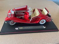 Side view of red Maisto model car with open hood showing engine detail, cream interior seats, and displayed on black base with name plaque.