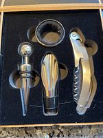 Close-up of four polished metallic wine accessories in black foam molded slots inside a wooden box.