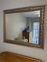 Framed wall mirror mounted on the wall with reflection showing part of room including furniture and glass items.