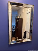 Front view of the rectangular wall mirror hung on a blue wall showing the beaded reflective frame and clear mirror surface. Part of a wooden cabinet and doorway reflected in the mirror.