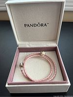 Pink braided leather wrap Pandora bracelet with silver Pandora clasp and one silver charm with embedded stones inside a white and pink Pandora box.