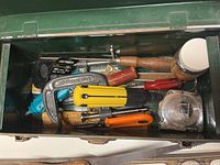 Opened green metal toolbox showing a variety of hand tools including pliers, utility knife, clamp, screwdriver, tape measure, and small woodworking supplies.