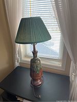 Full view of one pottery side table lamp on a black table in front of a window with curtains, showing the ceramic base with green and brown glaze and a pleated green fabric lampshade.