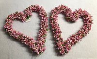 Two new heart-shaped wreaths decorated with small pink, white, and pastel-colored floral clusters and green leaves placed on a white surface.