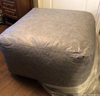 Grey pouf ottoman wrapped in plastic showing leather-look material with foam inside.