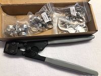 Photo showing Spinner clamp installation tool with grey handles positioned near plastic bags of PEX clamps in various sizes.