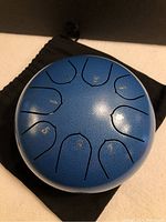 Overhead view of the metallic blue steel tongue drum showing 8 numbered tongue notes from 1 to 8 on the drum surface, resting on a black carry bag.