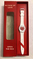 Red rectangular display box with 'IN CASE OF LOVE OPEN THE BOX' text, containing a new white-strap Swatch wristwatch with a heart design on the face.