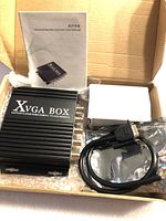 Top view of the open box showing XVGA Box converter device, cables, and user manual in non-English.