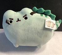 Front view of the pastel green Pusheenosaurus plush toy showing face and right side with character tag visible.