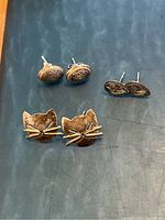 Front view of three pairs of silver stud earrings on dark surface, including cat face design and two patterned oval/rounded shapes.