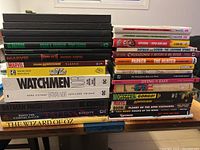 Stacked hardcover graphic novels including titles by Brian K. Vaughan, Marvel Comics, DC Comics, Dark Horse, and others on a table.