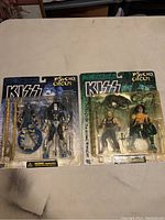 Two blister card packages featuring KISS Psycho Circus figures: Gene Simmons and Ace Frehley with their accessories, sealed and in good condition.