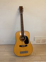 Full front view of Marlin M420 acoustic guitar standing upright against a wall on a wooden floor, showing overall guitar body and neck, natural wood color with a black pickguard.