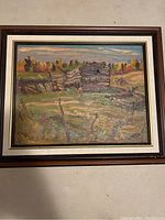 Frontal view of framed oil painting depicting old log farm building among fall-colored trees and open field under a cloudy sky.