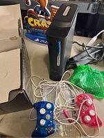 Xbox 360 Elite console, blue and pink wired Xbox 360 controllers, damaged original box, power supply cables visible.