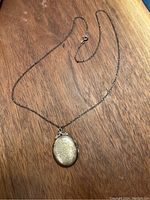 Full view of the silver oval locket pendant on chain laid on wood surface, showing front engraved design and chain length.