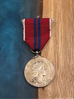 Front of silver round medal showing crowned left profile of Queen Elizabeth II with red ribbon having blue stripes and white edges.