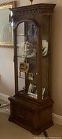 Full view of wooden display cabinet with glass shelves and interior lighting, filled with decorative items (not included). Shows front glass door, carved wood details, and base drawer with hardware.