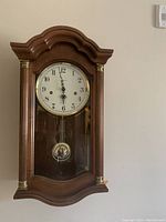 Front view of the Howard Miller wall clock showing cherry wood case, glass front, pendulum and clock face.