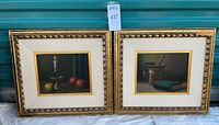 Two framed oil paintings showing a candle with apples and a mortar with book, each measuring approximately 18" x 17" with gold-tone frames and canvas matting.