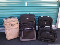 Five luggage and laptop bags displayed outside against a green corrugated metal door: a tan suitcase; black Pathfinder suitcase; black carry-on suitcase; black Samsonite laptop bag; black Delsey laptop bag