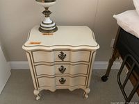 Full view of one nightstand showing cream finish, gold trim, three drawers, ornate hardware