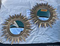 Pair of decorative sunburst mirrors laid flat on white cloth showing front reflective surfaces with textured gold tone frames surrounding circular convex glass.