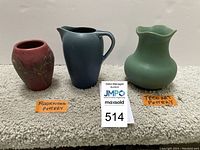 Three pottery pieces side by side on beige carpet: red Rookwood vase, blue ribbed pitcher, green TECO vase