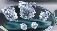 Three faceted Swarovski crystal swans in different sizes standing on an oval mirror base displaying the reflective qualities of the figurines.