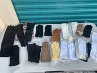 Photo showing a variety of gloves mostly leather in black, white, brown, and yellow with some fabric gloves and packages of new hosiery, also a pink and black scarf/head wrap shown underneath some gloves.
