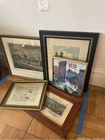 Five framed art pieces including vintage maps and cityscape prints on wood floor against wall.