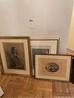 Set of three framed artworks leaning against a wall on a wooden parquet floor, including a classical portrait image and two other prints with matting.