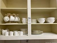 Full view of shelves showing the complete set of Aynsley dishes including tea cups on holder, broth bowls with saucers, sugar creamer, espresso cups, and dinner plates all in ivory color with gold trim.