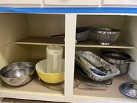 Photo of bakeware including mixing bowls, vintage Pyrex bowl, pitcher, aluminum pans, colander, and steamer basket inside a cabinet.