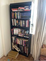 Front view of tall black bookcase filled with books, placed in front of window with curtain.