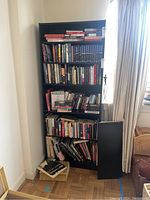 Full bookshelf view showing a black bookshelf filled with hardcover and paperback books, with some books stacked on the floor beside it.