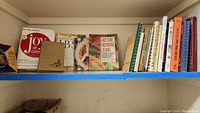 Shelf with 13 assorted cookbooks, including visible titles "Joy of Cooking", "Easy Down Home Cooking", and others mainly hardcover and spiral bound