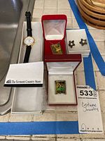 Photo showing wristwatch with white dial, black leather strap, rectangular pendants with nature images, and two pairs of star-shaped earrings in boxes.