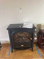 Full view of black Electralog compact electric stove with remote and paperwork on top, standing on hardwood floor with some blue tape marking under it.