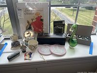 Photo of glass art and assorted decorative items on windowsill, including glass swan, black leather box, trinket boxes, wall plaques, snow globe music box, candle set, and decorative poster.