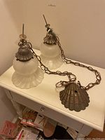 Two hanging light fixtures with frosted glass domes and brass seashell bases connected to metal chains with wiring, displayed on a white surface.
