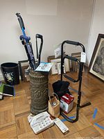 Photo showing several walking canes with distinct handles inside an ornate cane bucket, portable chair assist bar, ankle boot, blood pressure monitor, hearing aid batteries, and large button telephone on floor with framed pictures against wall in background.
